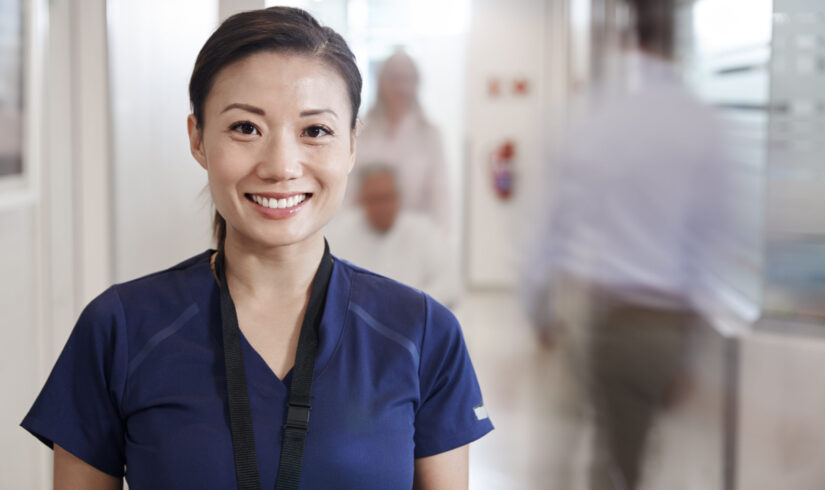 Portrait Of Smiling Female Nurse Wearing Scrubs In Busy Hospital Corridor